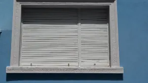 Une fenêtre blanche avec des volets fermés sur un mur bleu extérieur.