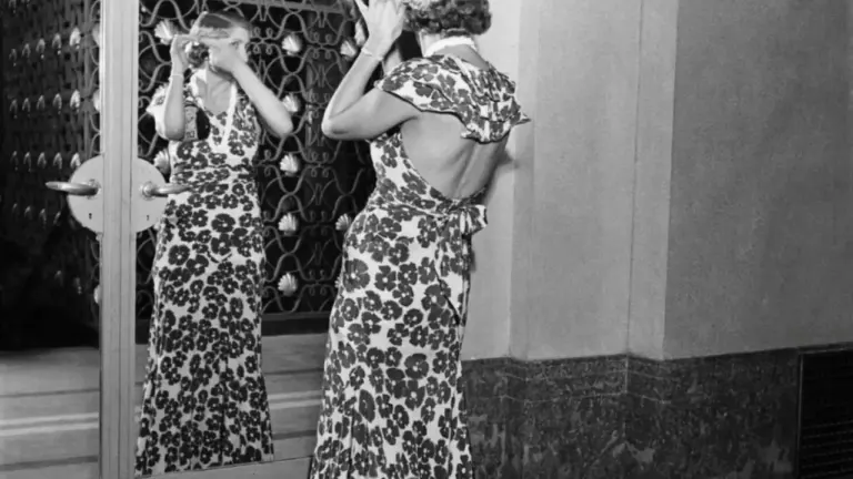 Une femme en robe longue fleurie se regarde dans un miroir décoré, ajustant sa coiffure.